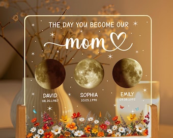 May include: A wooden frame with a clear acrylic insert featuring the text "The day you become our mom" with a heart symbol. Three crescent moons represent the dates of birth of three children with their names and birth dates below each moon. The frame is decorated with a floral border.