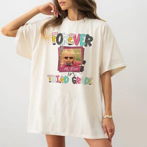 May include: An off-white short-sleeve t-shirt featuring a graphic print with the text FOREVER in colorful letters, a photograph of a child wearing sunglasses inside a pink frame, and the text Ms. Emma in THIRD GRADE with pencil icons.