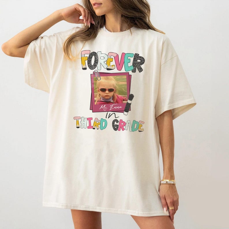 May include: An off-white short-sleeve t-shirt featuring a graphic print with the text FOREVER in colorful letters, a photograph of a child wearing sunglasses inside a pink frame, and the text Ms. Emma in THIRD GRADE with pencil icons.