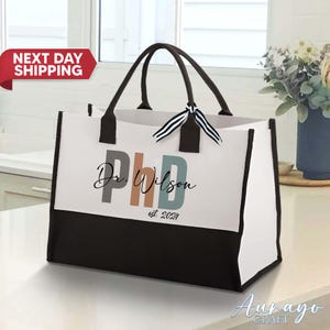 Pode incluir: Uma bolsa tote branca e preta com al&ccedil;as pretas. A bolsa tem um fundo preto e uma parte superior branca com o texto "Dr. Wilson PhD est. 2024" em letras coloridas.