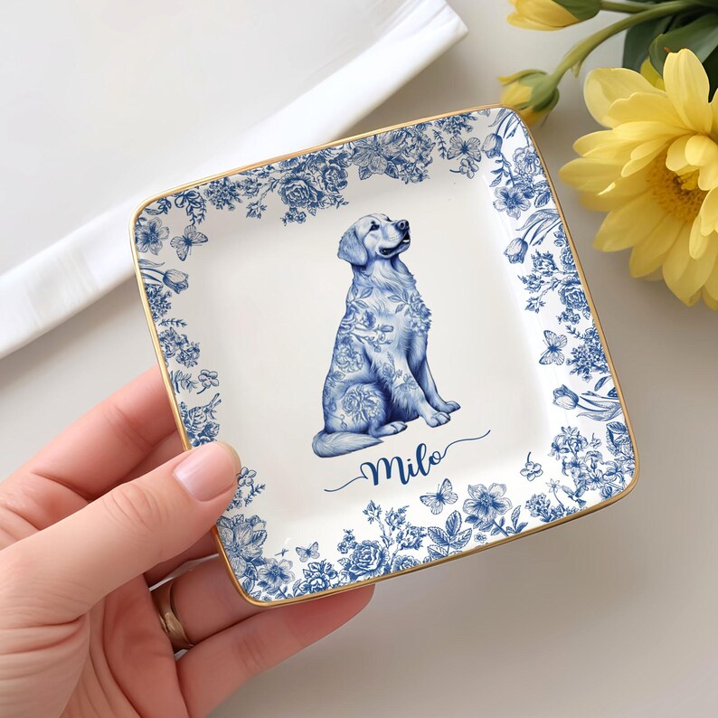 May include: Square ceramic trinket dish with a blue floral border and a gold rim. The center features a seated golden retriever illustration with floral patterns. The name "Milo" is written in cursive below the dog.