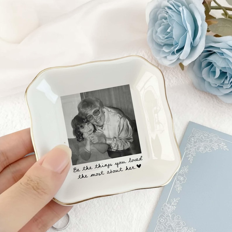 May include: A square white ceramic trinket dish with scalloped edges and a gold-toned rim. The centre features a black and white photograph of two people, with the text "Be the things you loved the most about her." printed in black script below.