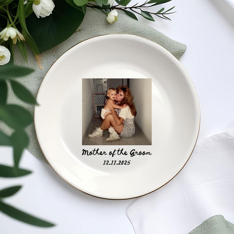 Op de afbeelding: Een witte keramische plaat met een gouden rand, met een vintage foto van een moeder en kind. De plaat is beschreven met "Mother of the Groom" en de datum 12.11.2025 in zwarte tekst. Decoratieve bloemelementen omringen de plaat.
