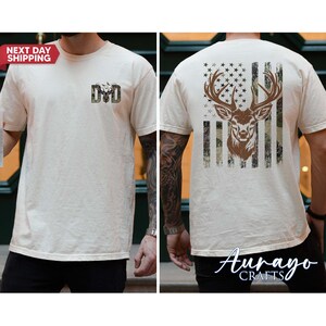 May include: Cream-colored t-shirt featuring a deer head design. The front displays a camouflage deer head and the letters "DD". The back showcases a deer head within a flag design. The shirt is short-sleeved.