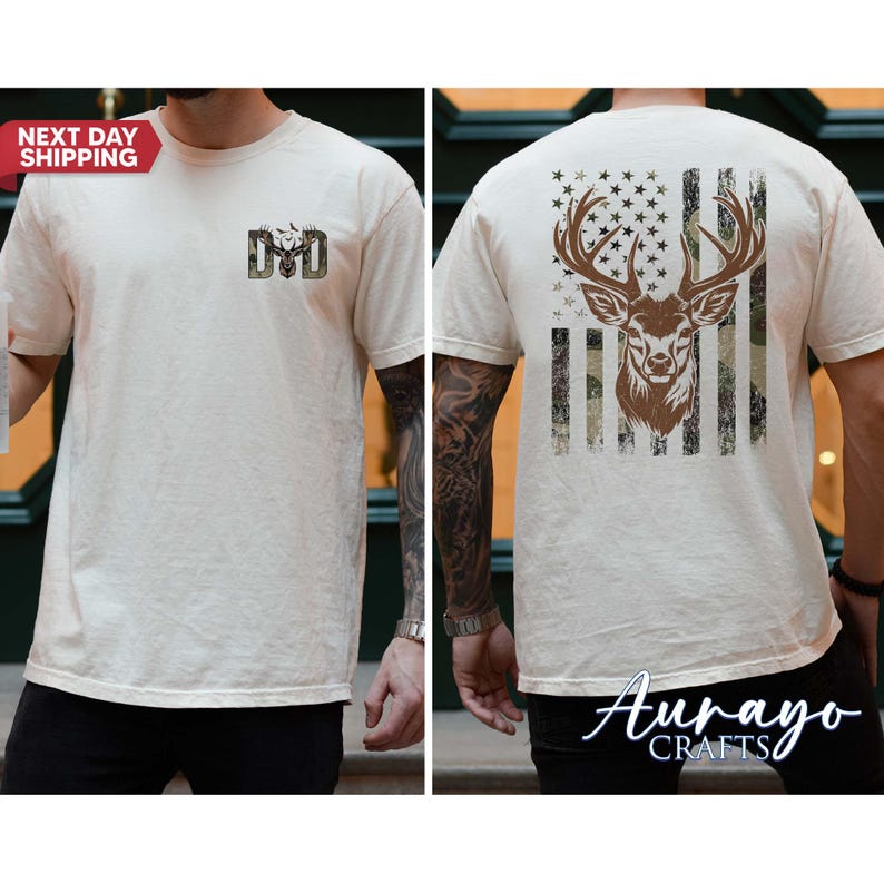 May include: Cream-colored t-shirt featuring a deer head design. The front displays a camouflage deer head and the letters "DD". The back showcases a deer head within a flag design. The shirt is short-sleeved.