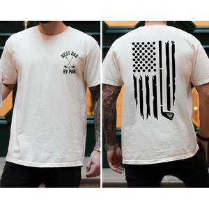 Peut inclure: Un t-shirt blanc avec un motif noir sur le devant qui dit "Best Dad By Par" avec deux clubs de golf crois&eacute;s. Le dos du t-shirt pr&eacute;sente un motif noir d'un club de golf et de balles de golf en forme de drapeau am&eacute;ricain.