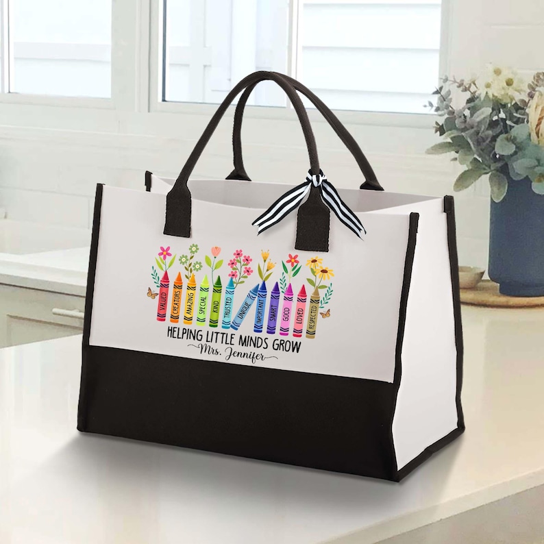 Puede incluir: Una bolsa de tela blanca y negra con un dise&ntilde;o colorido de l&aacute;pices de colores y flores. El texto en la bolsa dice "Helping Little Minds Grow - Mrs. Jennifer".