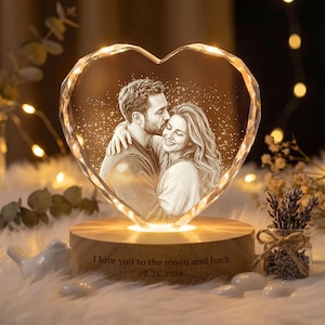 May include: A heart-shaped crystal plaque featuring an etched portrait of a couple, illuminated by a light base. The wooden base is engraved with the text, I love you to the moon and back, followed by the date 26.09.2026, in a clear font.
