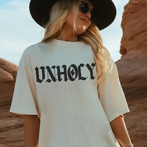 Op de afbeelding: Een wit T-shirt met het woord "UNHOLY" bedrukt in zwarte gotische letters. Het shirt wordt gedragen door een persoon met blond haar en een zwarte hoed.
