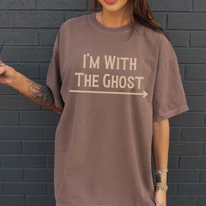 May include: A brown oversized t-shirt with the text "I'm with the ghost" and an arrow pointing to the right. The model is wearing white sneakers.