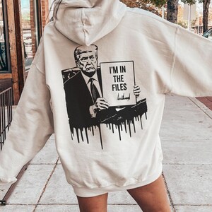Puede incluir: Sudadera con capucha color arena con un gráfico en la espalda. El gráfico es una imagen en blanco y negro de una persona sosteniendo un letrero que dice "I'M IN THE FILES". El gráfico tiene un efecto de pintura goteando. La sudadera se usa al aire libre.