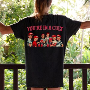 K&ouml;nnte beinhalten: Ein schwarzes T-Shirt mit einem grafischen Druck einer Gruppe von Kindern, die aus einem Brunnen trinken. Der Text "YOU'RE IN A CULT" ist &uuml;ber dem Grafikdruck gedruckt.