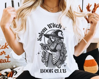 May include: White t-shirt with a black and white illustration of a witch reading a book. The text "Salem Witch Book Club" is printed on the shirt.