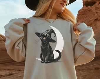 Puede incluir: Una sudadera gris con un gato negro que lleva un sombrero de bruja y está sentado en una luna creciente. El gato tiene ojos naranjas.