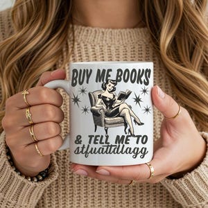 Op de afbeelding: Witte keramische mok met de tekst "BUY ME BOOKS & TELL ME TO stfuattdlaggg" en een vintage illustratie van een lezende vrouw. De mok heeft een wit handvat en wordt vastgehouden door een persoon die een beige trui draagt.