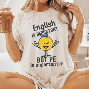 Puede incluir: Camiseta blanca con una cara sonriente amarilla que lleva una gorra de béisbol azul y sostiene un micrófono. El texto de la camiseta dice "English is important but PE is importanter".