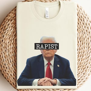 以下が含まれることがあります： ベージュのTシャツに、スーツを着た男性の白黒画像がプリントされています。男性の目は、「RAPIST」と書かれた黒いバーで覆われています。