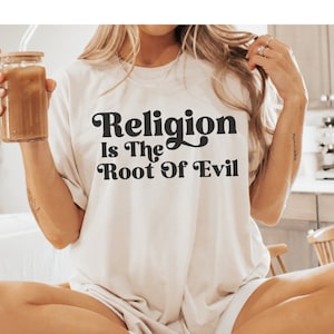 Può includere: Una maglietta bianca con la scritta "Religion is the Root of Evil" stampata in nero con un font in stile retrò.