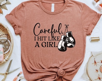 Peut inclure: Un t-shirt rose avec le texte "Careful, I hit like a girl" et une paire de gants de boxe suspendus à une ficelle.