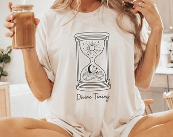 May include: A white t-shirt with a black line drawing of an hourglass with a sun, moon, and waves inside. The text "Divine Timing" is below the hourglass.