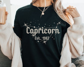 Könnte beinhalten: Ein schwarzes T-Shirt mit einem goldenen Glitzer-Design der Sternbilder des Steinbocks und dem Text "Capricorn Est. 1987".