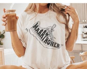 Peut inclure: T-shirt blanc avec une illustration noir et blanc d'une chaussure de course avec le mot "MARATHONER" écrit dessus. Le t-shirt est porté par une personne assise sur une surface en bois. La personne porte des chaussettes blanches avec des rayures noires.