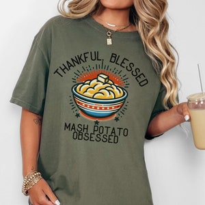 Puede incluir: Camiseta verde salvia con un gráfico de puré de patatas en un cuenco, una nuez de mantequilla y las palabras "Thankful, Blessed, Mash Potato Obsessed". La camiseta tiene un corte holgado y mangas cortas. La palabra "SAGE" está impresa abajo a la derecha.