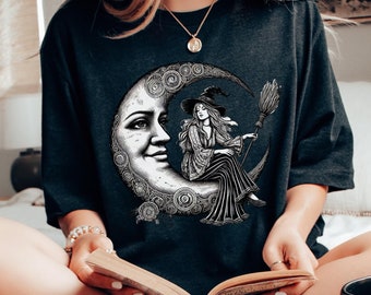 May include: A black t-shirt with a graphic of a witch sitting on a crescent moon. The witch is wearing a pointed hat and holding a broom. The moon is decorated with swirls and the graphic is in black and white.