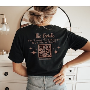 May include: Black t-shirt with the text "The Bride I'm Tying The Knott Buy Me A Shot" in rose gold lettering. A QR code is printed below the text with the words "@YOUR VENMO" underneath.
