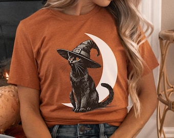 May include: A brown t-shirt with a black cat wearing a witch's hat and sitting on a crescent moon. The cat has orange eyes.
