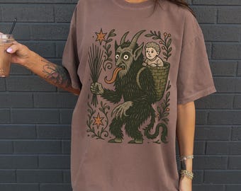 May include: Espresso-colored t-shirt featuring a whimsical illustration of a horned creature carrying a child in a basket. The design includes stars and foliage. The shirt has short sleeves and a relaxed fit.