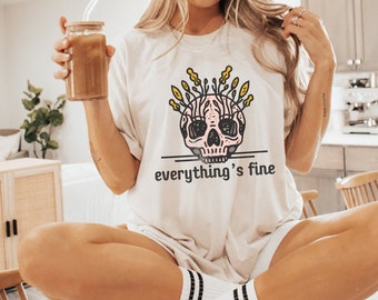 Può includere: Una maglietta bianca con un grafico di teschio rosa con fiori gialli che crescono dalla cima del teschio. Il testo "everything's fine" è stampato sotto il teschio. La modella indossa la maglietta e siede su una superficie bianca.