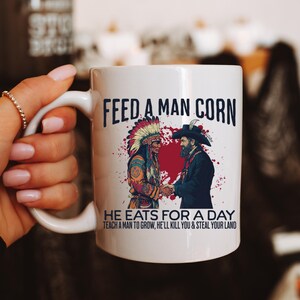Könnte beinhalten: Weiße Keramik-Tasse mit dem Text "FEED A MAN CORN" und einer Illustration von zwei Männern, die sich die Hände schütteln. Die Tasse enthält auch den Text "HE EATS FOR A DAY TEACH A MAN TO GROW, HE'LL KILL YOU & STEAL YOUR LAND."