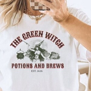 May include: White t-shirt featuring a green witch graphic and the text "The Green Witch Potions and Brews Est. 1626".