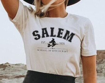 Puede incluir: Una mujer con una camiseta blanca con el texto "Salem School of Witchcraft Est. 1626" y una falda negra. Lleva un sombrero negro y está de pie en un paisaje desértico.