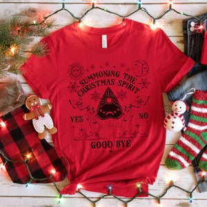 May include: Red t-shirt with a black graphic of a Ouija board with the text "Summoning the Christmas Spirit" and "Good Bye" on a white background.