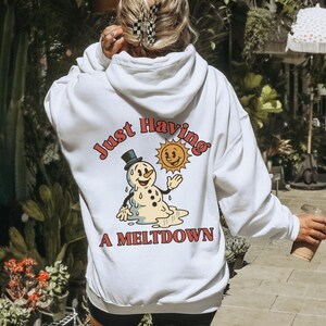 May include: White hoodie with a graphic on the back featuring a melting snowman wearing a top hat and a smiling sun. The text reads "Just Having A Meltdown" in a retro font. The hoodie has a hood and long sleeves.
