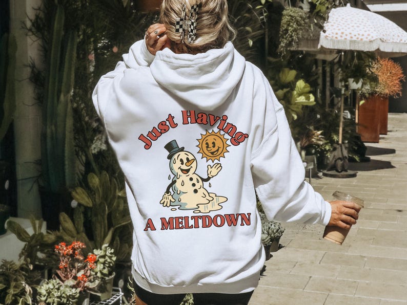 May include: White hoodie with a graphic on the back featuring a melting snowman wearing a top hat and a smiling sun. The text reads "Just Having A Meltdown" in a retro font. The hoodie has a hood and long sleeves.
