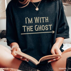 May include: A woman wearing a black t-shirt with the text "I'm with the ghost" in gold lettering and an arrow pointing to the right. She is sitting on a bed and reading a book.