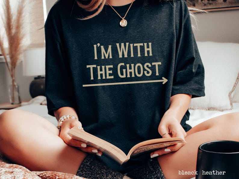 May include: A woman wearing a black t-shirt with the text "I'm with the ghost" in gold lettering and an arrow pointing to the right. She is sitting on a bed and reading a book.