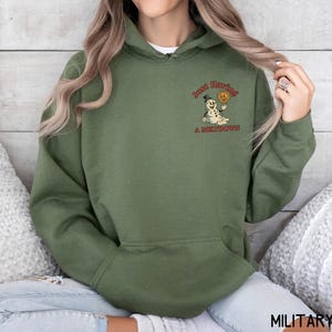 May include: Military green hoodie with a front pocket and drawstring hood. The hoodie features a small graphic on the upper left chest of a snowman holding a smiley face with the text "Just Having A Meltdown".