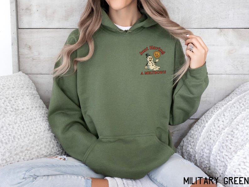 May include: Military green hoodie with a front pocket and drawstring hood. The hoodie features a small graphic on the upper left chest of a snowman holding a smiley face with the text "Just Having A Meltdown".