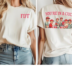 K&ouml;nnte beinhalten: Ein wei&szlig;es T-Shirt mit dem Text "FDT" auf der Vorderseite und "YOU'RE IN A CULT" auf der R&uuml;ckseite. Das Design auf der R&uuml;ckseite zeigt eine Gruppe von Cartoon-Kindern, die aus Gl&auml;sern trinken.