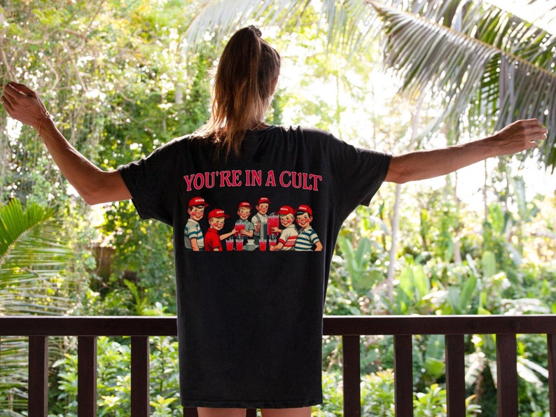 K&ouml;nnte beinhalten: Ein schwarzes T-Shirt mit einem grafischen Druck einer Gruppe von Kindern, die aus einem Brunnen trinken. Der Text "YOU'RE IN A CULT" ist &uuml;ber dem Grafikdruck gedruckt.