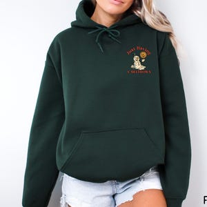 May include: Forest green hoodie with a front pocket and drawstring hood. The upper left chest features a graphic of a snowman holding a sun with the text "Just Having A Meltdown" in orange. The word "FOREST" is printed in the bottom right corner.