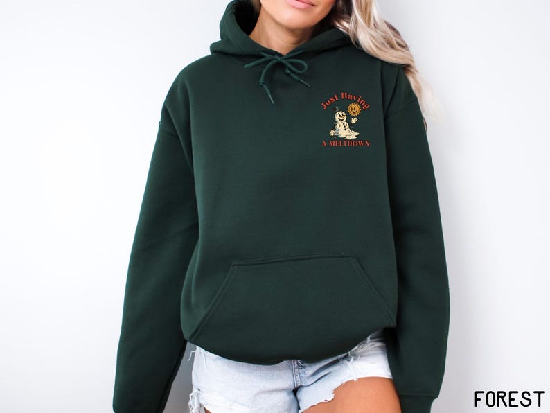 May include: Forest green hoodie with a front pocket and drawstring hood. The upper left chest features a graphic of a snowman holding a sun with the text "Just Having A Meltdown" in orange. The word "FOREST" is printed in the bottom right corner.