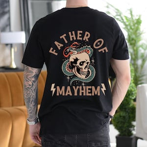 以下が含まれることがあります： 骸骨に蛇が巻き付いているグラフィックプリントが施された黒いTシャツ。骸骨の下に「Father of Mayhem」という文字がプリントされています。