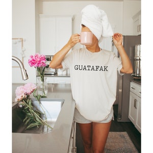 May include: A woman wearing a white t-shirt with the text "GUATAFAK" printed on it. She is holding a pink mug and has a white towel wrapped around her head.