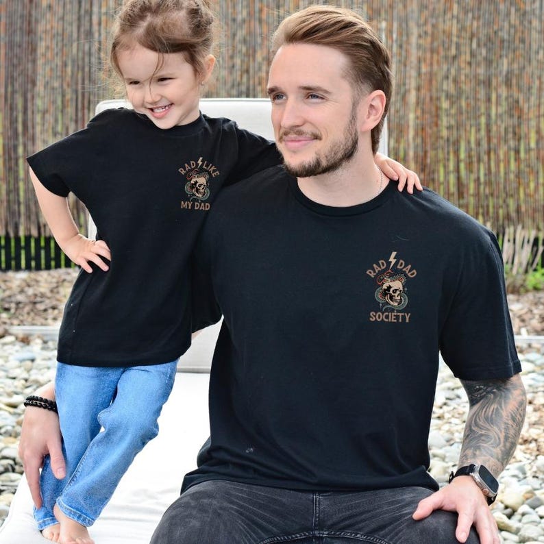 May include: A father and daughter wearing matching black t-shirts with a skull and snake graphic. The father's shirt says "Rad Dad Society" and the daughter's shirt says "Rad Like My Dad".
