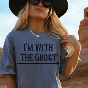 May include: A woman wearing a blue t-shirt with the text "I'm with the ghost" and an arrow pointing to the right. She is wearing a black hat and sunglasses. The background is a desert landscape.